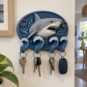May include: A wall-mounted key holder featuring a three-dimensional grey shark emerging from blue ocean waves. The oval-shaped plaque includes four integrated hooks, each holding a metal keyring with various keys and a black car key fob.