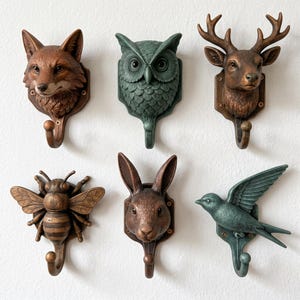 Peut inclure: Un ensemble de six patères murales représentant des têtes et des figures d'animaux, dont un renard, un hibou, un cerf, une abeille, un lapin et un oiseau. Les crochets présentent des finitions en bronze métallique et en tons vert sourd.