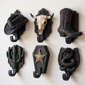 May include: A set of six wall-mounted hooks featuring western-themed designs including a black cowboy hat with barbed wire, a white bull skull, a black and red cowboy boot, a green cactus, a black coffin with a gold star, and a black coiled snake.