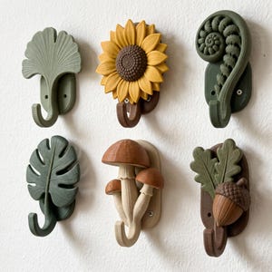 May include: A set of six wall-mounted hooks featuring nature-inspired designs including a ginkgo leaf, a yellow sunflower, a green fern frond, a monstera leaf, a cluster of three brown mushrooms, and an acorn with oak leaves, all mounted with screws.