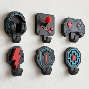 May include: A set of six black wall hooks featuring gaming-themed designs including a headset, a joystick, a game controller, a lightning bolt, a shield, and a geometric emblem, all accented with vibrant red and bright blue color details.
