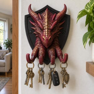 Puede incluir: Un soporte de pared para llaves con una cabeza de dragón roja esculpida sobre una placa negra en forma de escudo. El dragón tiene cuatro garras curvadas hacia abajo que sirven de ganchos, cada una con una anilla metálica con varias llaves.