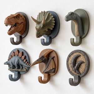 Könnte beinhalten: Ein Set aus sechs Wandhaken mit Dinosaurier-Motiven. Die Kollektion umfasst einen T-Rex-Kopf, einen Triceratops-Kopf, einen Langhals-Dinosaurier-Kopf, einen Stegosaurus, einen Pterodaktylus und eine versteinerte Klaue auf runden Sockeln.
