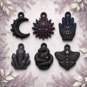 Pode incluir: Um conjunto de seis pingentes pretos com designs em relevo, incluindo uma lua crescente, um rosto de sol, uma mão hamsa com um olho, um aglomerado de cristais, uma cobra enrolada com estrelas e uma mariposa com uma estrela, cada um com um aro.