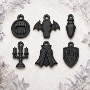 Puede incluir: Un conjunto de seis dijes de color negro mate con motivos góticos que incluyen un par de colmillos de vampiro, un ataúd con alas de murciélago, un vial puntiagudo, un candelabro de tres brazos, una capa y un escudo con un castillo detallado.