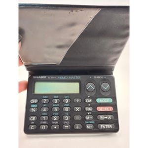 Könnte beinhalten: Ein schwarzer Sharp EL-6061 Memo Master elektronischer Organizer mit aufklappbarem Deckel. Das Gerät verfügt über ein digitales Display, eine vollständige numerische Tastatur mit integrierten Buchstaben sowie Funktionstasten für Mode, Secret und Enter.