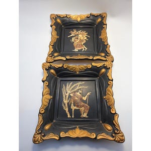 Puede incluir: Dos platos decorativos cuadrados en negro y oro con bordes ornamentados en relieve y detalles dorados. El centro de cada plato muestra una ilustración detallada en tono dorado de una figura clásica, una de pie y otra sentada bajo un árbol.