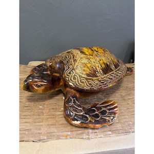 Puede incluir: Una figura de tortuga marina de cerámica con un caparazón brillante en tonos marrón y amarillo, decorado con un borde en relieve de nudos celtas. La cabeza y las aletas tienen un esmalte marrón oscuro con texturas de escamas visibles.