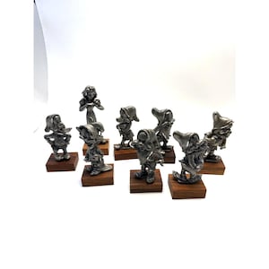 Op de afbeelding: Een verzameling van acht zilverkleurige metalen beeldjes die personages uit een klassiek sprookje voorstellen. Elk beeldje staat op een kleine, vierkante voet van donkerbruin hout. De set bevat één groter centraal figuur.