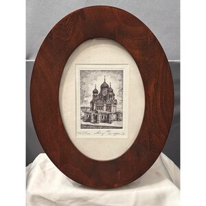 May include: An oval-shaped dark wood frame containing a black and white etching of a cathedral with multiple domes. The artwork is set against a cream-colored mat board and features handwritten text at the bottom that reads Tallinn Alexander 1/10 copper.