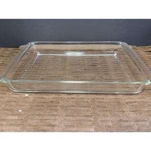 Puede incluir: Una fuente de horno rectangular de vidrio transparente con una superficie lisa y esquinas ligeramente redondeadas. El centro de la base presenta el texto Pyrex en relieve, indicando la marca de este utensilio de cocina para horno.