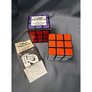 May include: A 3x3 cube puzzle with orange, white, and black plastic segments. The set includes a black cardboard box with purple accents labelled CUBE PUZZLE and an instruction manual featuring a diagram and text describing the logic challenge.