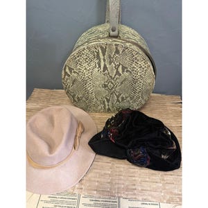 May include: A round, structured case with a snakeskin pattern in shades of beige and brown, featuring a top handle. Beside it sits a light beige felt fedora hat with a matching band and a black velvet hat adorned with colourful floral accents.