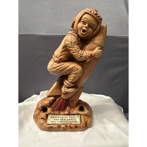May include: A tan-colored figurine of a child wearing a flight helmet and suit, riding a rocket ship. The figure has a red cross emblem on the sleeve. A plaque at the base reads: COMMUNITY CHEST AND RED CROSS 1955 CAMPAIGN AWARD. The rocket has red flames.