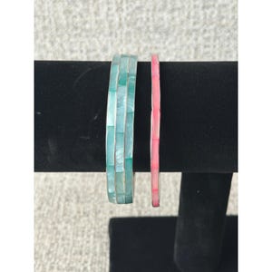 May include: Two bangle bracelets are displayed side by side. The wider bracelet features a mosaic of rectangular, iridescent teal-green shell tiles. The thinner bracelet is composed of a single row of rectangular, iridescent pink shell tiles.