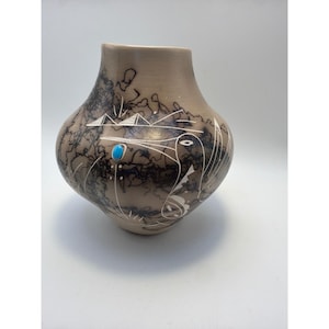 May include: A tan ceramic vase with a rounded body and narrow neck, featuring black horsehair-style smoke markings, white etched geometric and bird-like patterns, and a single small, oval, bright blue turquoise stone inlaid on the front.