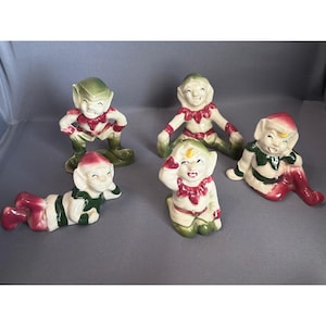 Peut inclure: Un ensemble de cinq figurines d'elfes en céramique dans des poses variées. Chaque elfe présente un corps de couleur crème avec des accents verts et rouges sur ses vêtements, son chapeau et ses chaussures. Les visages sont peints avec soin.