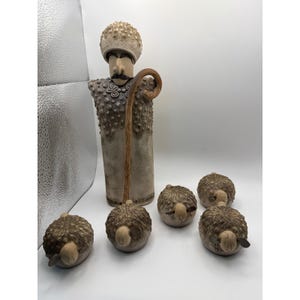 Puede incluir: Un conjunto de esculturas de cerámica que incluye una figura alta con un sombrero texturizado y una túnica estampada, sosteniendo un bastón curvo. La figura está acompañada por cuatro pequeñas figuras de ovejas de cerámica redondas.