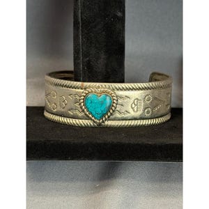 May include: A silver-toned metal cuff bracelet featuring a central heart-shaped turquoise stone with a rope-style border. The band is decorated with stamped geometric patterns, including sunbursts and arrow-like motifs, and has a twisted rope edge.