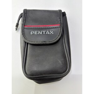 May include: A black rectangular camera case with a fold-over flap closure. The word PENTAX is embossed in gray on the front flap, which features a thin horizontal red line detail. The case has a small black strap attached to the side.