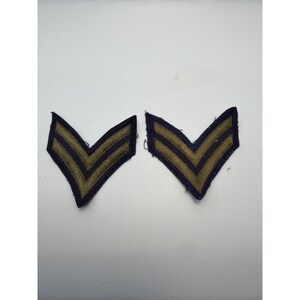 May include: A pair of embroidered military-style chevron patches featuring two parallel olive green stripes bordered by dark navy blue stitching. These V-shaped insignia patches are designed with a pointed bottom edge and a flat top edge.