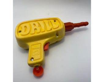 May include: A yellow plastic toy drill with the word DRILL embossed on the side in large capital letters. The tool features a red trigger, a red drill bit extending from the front, and a small red circular component attached to the bottom handle.