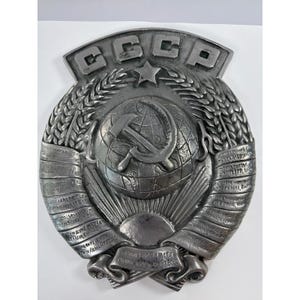 May include: A silver-toned metal wall plaque featuring the Soviet Union emblem. The design includes a globe with a hammer and sickle, surrounded by wheat stalks, a five-pointed star, and the Cyrillic text CCCP at the top and ribbons below.