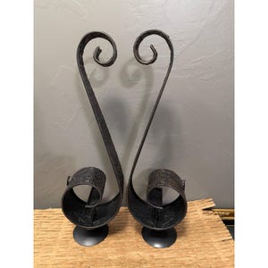 May include: A pair of dark brown metal candle holders featuring a tall, slender, S-shaped curved design that spirals at the top and bottom. The metal surface is textured with an embossed floral pattern, and each holder stands on a circular base.