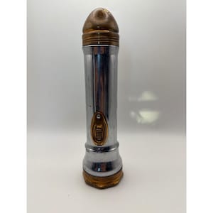 May include: A vintage-style cylindrical flashlight featuring a polished chrome body with a rounded, brass-toned top cap and a matching brass-toned base. The center of the handle includes an oval-shaped brass switch mechanism for operation.