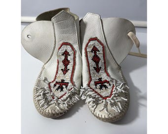 May include: A pair of white leather moccasins featuring intricate beadwork on the vamps. The beadwork displays a geometric pattern in red, black, and white, accented with white beaded fringe at the toes and side lace closures.