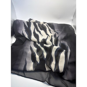 Könnte beinhalten: Ein leichtes, halbtransparentes quadratisches Tuch mit einem markanten, abstrakten Tigerstreifenmuster in den Farben Schwarz, Anthrazit und Cremeweiß. Der Stoff hat eine glatte, fließende Textur mit einem einfarbigen schwarzen Rand.