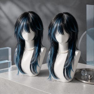 May include: Two identical wigs displayed on white mannequin heads. Each wig features long, layered black hair with prominent streaks of vibrant blue throughout. The hair is styled with a shaggy, textured cut, including bangs that frame the face.
