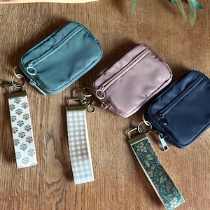 May include: Three rectangular zippered pouches in sage green, dusty rose, and black. Each pouch features a matching fabric wristlet strap attached by a metal ring. The straps display distinct patterns: floral, beige gingham, and botanical.