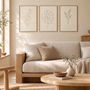 May include: Three framed wall art prints featuring black line drawings of botanical elements including a leafy branch, a monstera leaf, and pampas grass in a vase, displayed in light wood frames on a neutral wall above a beige sofa.