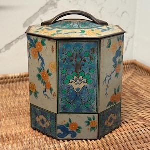 May include: An octagonal metal canister featuring a cream-colored background with orange floral patterns and blue decorative panels. The lid includes a dark metal handle, and the overall design displays intricate botanical motifs in blue, green, and orange.