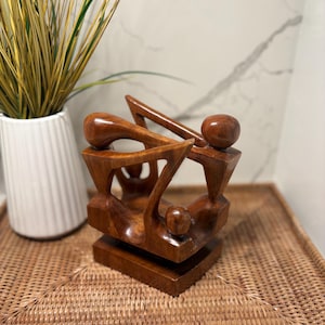 May include: A brown wooden sculpture featuring three stylized figures with smooth, rounded heads and angular bodies connected in a circular formation, all mounted on a rectangular wooden base with a polished, glossy finish.
