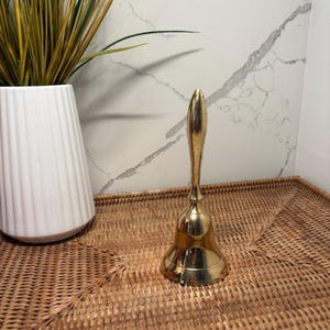 May include: A polished brass hand bell featuring a smooth, elongated handle and a flared base. The metal surface is reflective and gold-toned, showcasing a classic bell shape designed for ringing. The object stands upright on a woven surface.