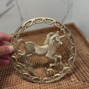 May include: A circular gold-toned metal wall plaque featuring a relief sculpture of a unicorn in the center. The unicorn is depicted in a standing position, surrounded by an ornate, openwork border with a repeating oval pattern throughout the frame.