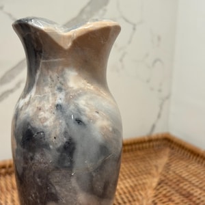 May include: A stone vase featuring a smooth, polished surface with a marbled pattern in shades of grey, black, white, and tan. The vase has a narrow neck that flares outward at the top rim, creating a distinct, petal-like opening shape.