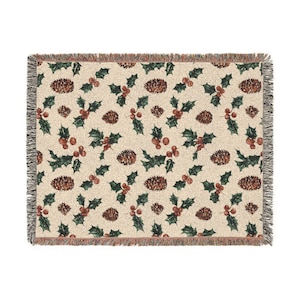 May include: A woven throw blanket with a cream-colored background featuring a repeating pattern of dark green holly leaves, bright red berries, and brown pinecones. The blanket has a textured fabric finish and frayed edges on all four sides.