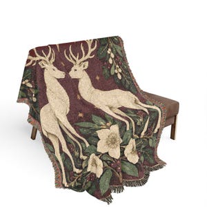 May include: A woven throw blanket featuring a deep burgundy background with two cream-colored deer facing each other. The design includes green pine branches, white flowers, and small gold stars. The blanket has a fringed edge on all sides.