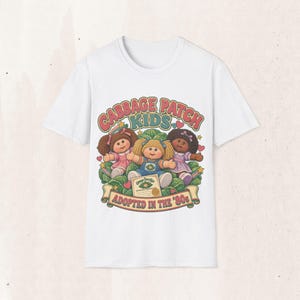 May include: A white short-sleeved t-shirt featuring a graphic of three Cabbage Patch Kids dolls sitting in a cabbage patch. The text reads Cabbage Patch Kids and Adopted in the '80s, with an adoption certificate illustration included in the design.