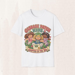 May include: A white short-sleeved t-shirt featuring a graphic of three Cabbage Patch Kids dolls sitting in a cabbage patch. The text reads Cabbage Patch Kids and Adopted in the '80s, with an adoption certificate illustration included in the design.