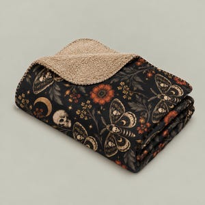 May include: A folded throw blanket with a black background featuring a pattern of moths with skull markings, human skulls, crescent moons, stars, and orange flowers. The reverse side of the blanket is made of a textured, tan-colored fleece material.