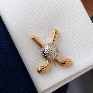 Puede incluir: Un gemelo de metal dorado que presenta dos palos de golf cruzados detrás de una pelota de golf plateada y texturizada. El accesorio está sujeto a un puño de tela blanca con un tejido visible, que contrasta con un fondo de tela azul oscuro.