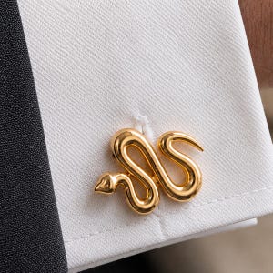 Può includere: Un gemello in metallo color oro a forma di serpente avvolto con una finitura liscia e lucida. Il serpente è raffigurato in una postura sinuosa a forma di S con una testa definita e una coda affusolata, fissato nell'asola di un polsino bianco.