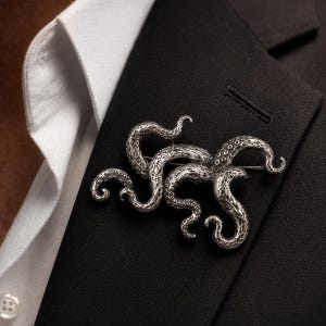 Peut inclure: Une broche en forme de tentacules de pieuvre argentée présentant une surface texturée très détaillée avec des ventouses visibles. L'accessoire métallique est épinglé sur le revers d'un blazer noir porté sur une chemise blanche à col boutonné.