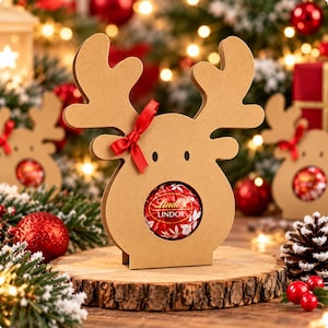May include: A tan cardboard reindeer-shaped holder featuring a circular cutout in the center that displays a round red Lindt Lindor chocolate truffle. The reindeer has two small black eyes and a small red ribbon tied to one of its antlers.