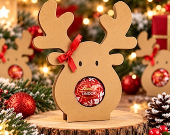 May include: A tan cardboard reindeer-shaped holder featuring a circular cutout in the center that displays a round red Lindt Lindor chocolate truffle. The reindeer has two small black eyes and a small red ribbon tied to one of its antlers.