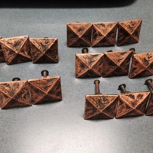 May include: A collection of twelve square cabinet knobs with a pyramid-shaped top and a textured, weathered copper finish. Each knob features a central point and a metal mounting screw extending from the back, arranged in four groups of three.